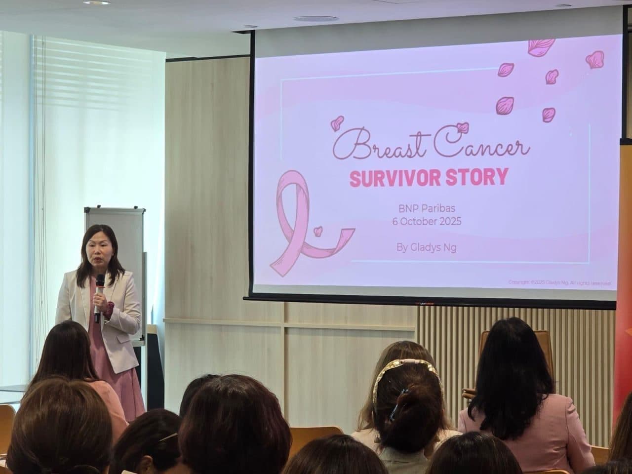 Gladys speaking into a microphone beside a screen titled “Breast Cancer Survivor Story”